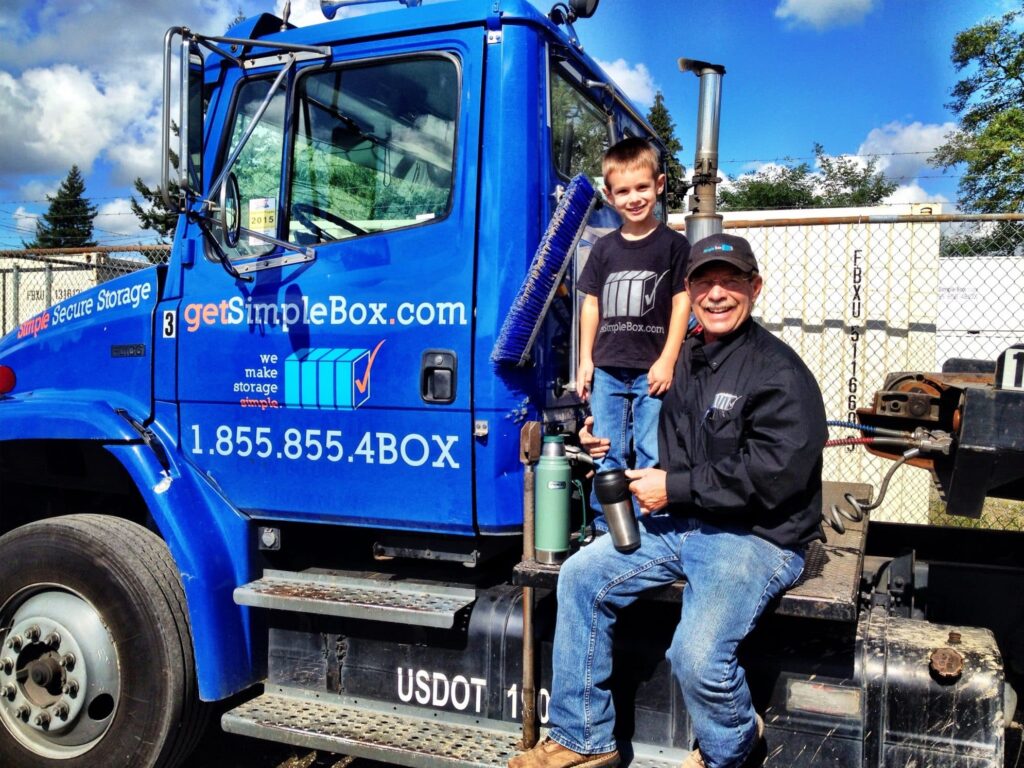 A boy and his grandfather sitting near a delivery truck. The truck is blue. The people are wearing black shirts and blue jeans.