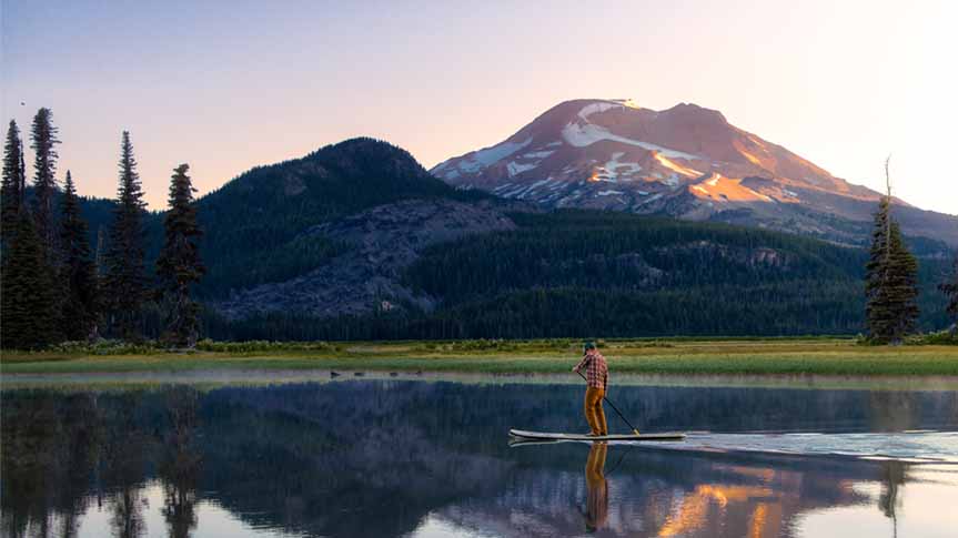 Picturesque view of mountains behind a man paddle boarding on a lake in Oregon.