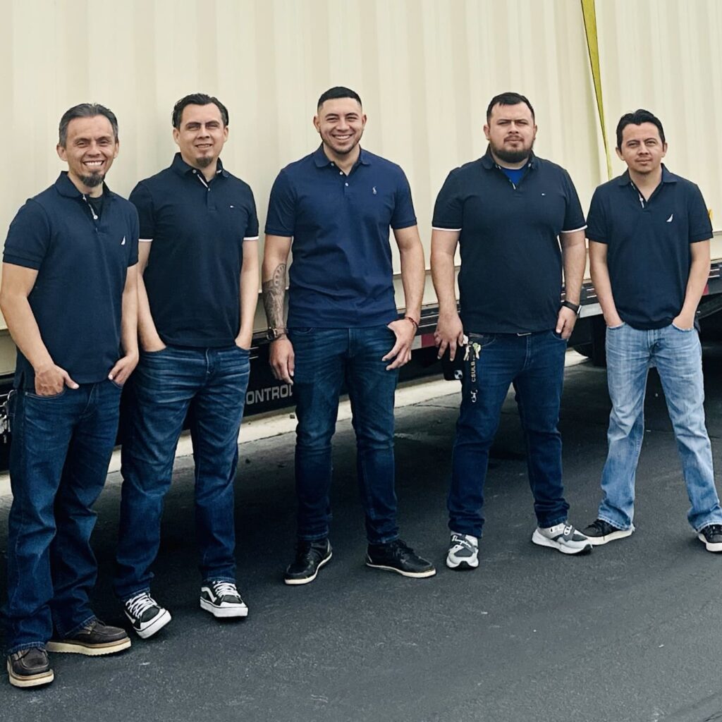 Five brothers standing in front of a Shipping Container. The men are wearing blue shirts and pants. The container is tan in color.