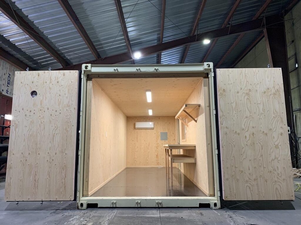 A shipping container that has been converted into a custom workspace with lights and work bench. the Container is beige and the walls are lined with plywood.