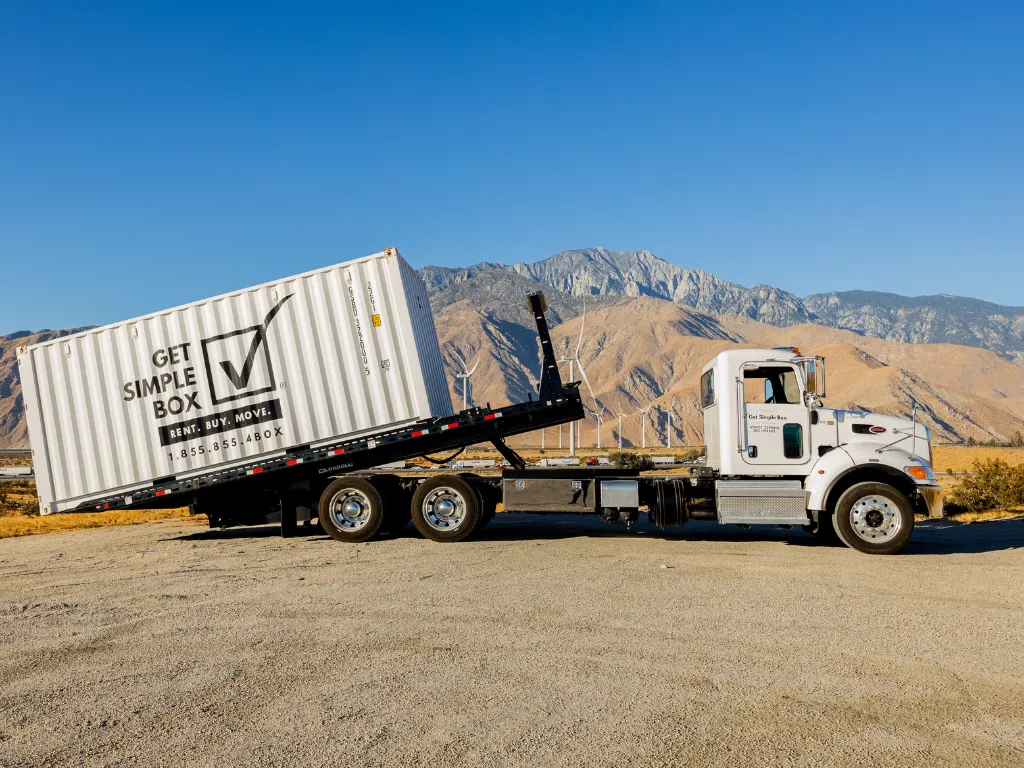 White delivery truck hauling a shipping container on a tilt-bed trailer in a desert lot with mountains in the background